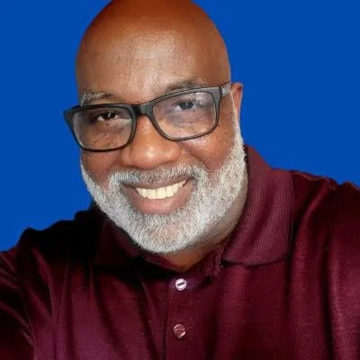 Professional portrait of DJ Travis Gales wearing glasses and a burgundy polo shirt against a royal-blue background